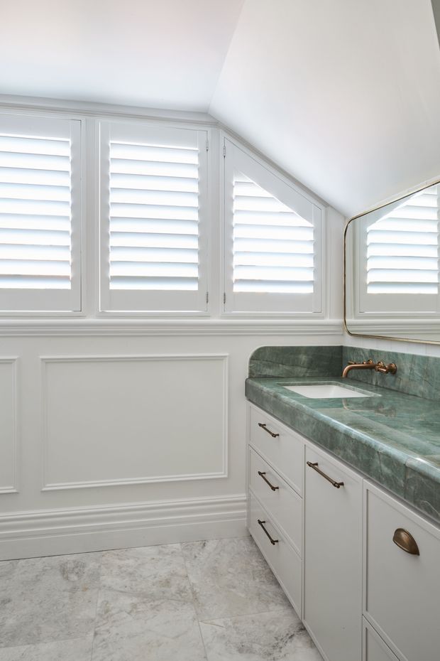 Bright, refined bathroom vanity area with white plantation shutters, detailed white wall panelling, a green veined stone benchtop and integrated basin, antique brass wall-mounted tapware and hardware, and pale stone-look floor tiles. A large framed mirror reflects the shutters and vanity.