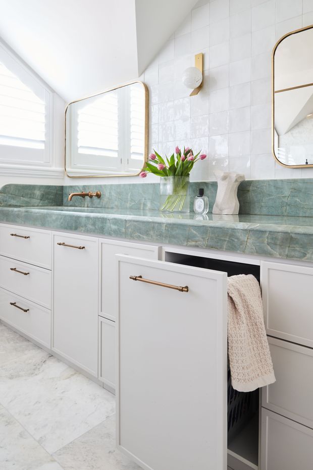 Bright, refined bathroom vanity with white paneled cabinetry, brass hardware, a green veined stone benchtop and matching backsplash, white tiled walls, arched mirrors, and a pull-out laundry hamper with towel storage. Natural light enters through a shuttered window, creating a fresh traditional-contemporary feel.