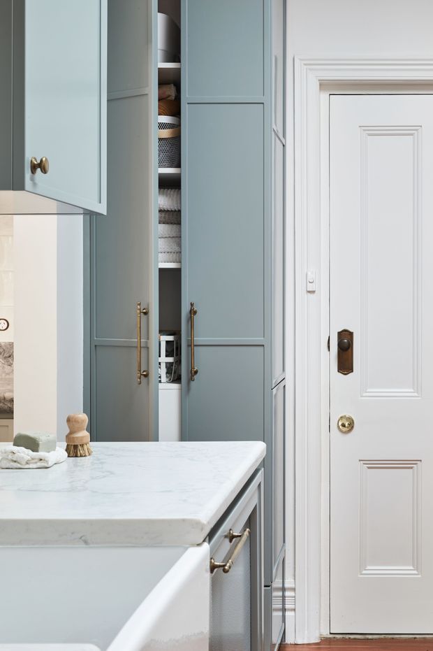 A refined, compact bathroom vanity area with pale blue-grey built-in cabinetry, brass hardware, and a white marble countertop. A white paneled interior door with ornate traditional moulding stands beside the storage cupboards, while neatly arranged towels and bathroom accessories are visible in the open cabinet.
