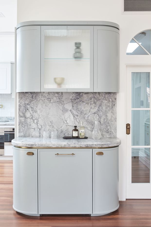 A refined built-in kitchen drinks cabinet or butler’s pantry feature with curved pale grey cabinetry, a glass-front display cupboard, dramatic veined stone splashback and matching stone benchtop. Brass hardware, glassware and bottles provide subtle accents. The cabinet sits against a light interior with timber flooring and glazed French-style doors visible to the side.
