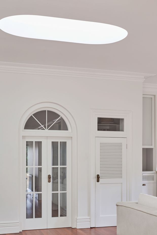 A refined white interior featuring a glazed French-style double door with an arched fanlight, a louvered built-in cupboard door, ornate wall and ceiling cornices, and polished timber flooring. A large oval flush-mount ceiling light and part of a pale sofa are visible, creating a bright, elegant transitional space.