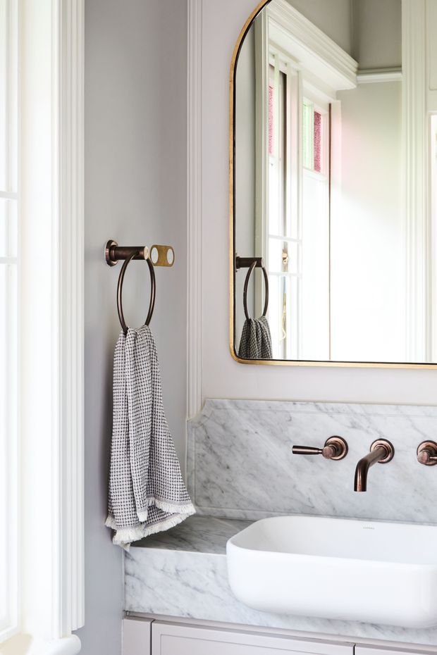 A bright, refined bathroom vanity with a white vessel basin set into a veined marble benchtop and splashback. The wall-mounted rose-gold/bronze tapware and matching towel ring sit against a soft grey wall with detailed white architectural mouldings. A large rounded-rectangle mirror with a warm metallic frame reflects tall windows and decorative interior trim.