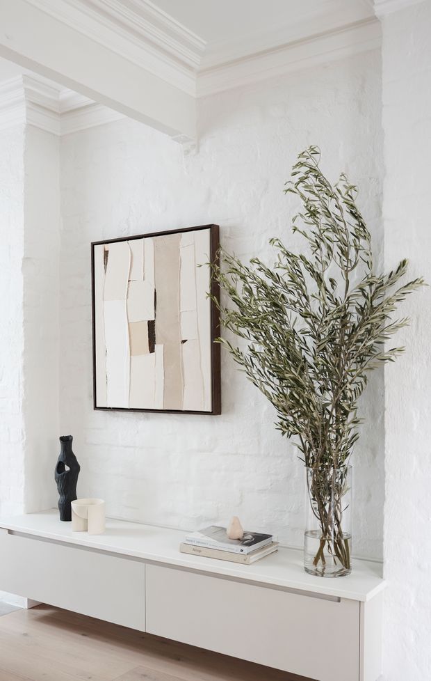 A bright, minimalist interior vignette featuring a white-painted textured brick wall, ornate white ceiling cornices, and a long floating white built-in cabinet. A framed abstract artwork, sculptural black vase, small ceramic vessels, stacked books, and a tall glass vase of olive branches add restrained decorative detail. Pale timber flooring and predominantly white finishes create an airy, refined atmosphere.