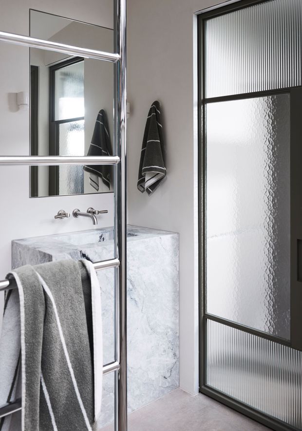 A compact contemporary bathroom viewed through a chrome-framed glass shower enclosure. The room features a white wall-mounted basin with a pale grey veined stone surround, wall-mounted chrome tapware, a large mirror, grey striped towels, and a black-framed textured and ribbed glass door or partition. The palette is monochromatic grey, white, and black with polished metal accents.