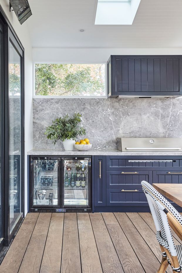 A compact, bright kitchen with deep navy-blue paneled cabinetry, brass hardware, a grey veined stone backsplash, and a stainless-steel rangehood. The space includes a glass-front beverage refrigerator, timber-look flooring, a skylight, and large windows overlooking greenery. A woven dining chair and timber tabletop are visible in the foreground.