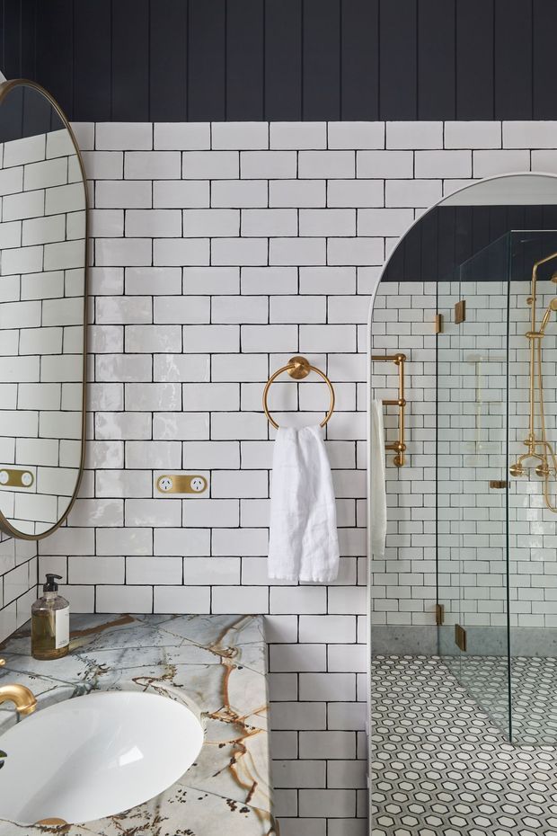 A refined bathroom featuring white subway-tile walls with dark grout, a marble vanity countertop and white vessel basin, arched mirrors, and a glass-enclosed shower. Brass and gold-toned fixtures, towel hardware, and trim add warmth, while patterned hexagonal floor tiles continue into the shower area.