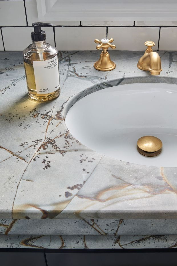 Close-up view of a bathroom vanity featuring a white oval undermount basin set into a dramatic veined stone countertop, with warm brass cross-handle tapware and matching drain stopper. White subway wall tiles and a soap dispenser are visible.