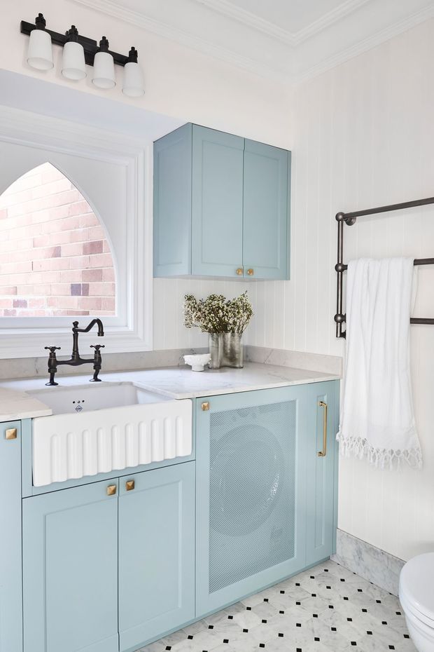 Bright, refined bathroom featuring pale blue built-in cabinetry, a white farmhouse-style apron-front basin, white marble-look benchtop, black traditional tapware and towel rail, and a patterned white-and-black tiled floor. White vertical wall panelling, decorative ceiling mouldings, and an arched window with exposed brick visible beyond add classic architectural character.