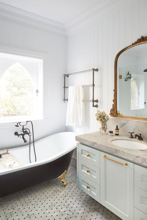 Bright, elegant traditional bathroom featuring white vertical wall panelling, ornate cornice detailing, a freestanding clawfoot bathtub with dark exterior, polished metal bath fittings, patterned floor tiles, and a pale blue-grey vanity with gold hardware. A marble-look benchtop, integrated basin, and large ornate gold-framed mirror complete the refined design.
