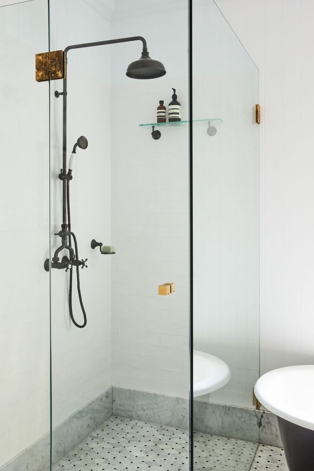 Bright bathroom with a frameless glass walk-in shower, dark bronze/black shower fixtures, white subway wall tiles, patterned light floor tiles, and a large wall mirror reflecting the curved white basin and toilet. Brass and dark metal accents add contrast to the predominantly white scheme.
