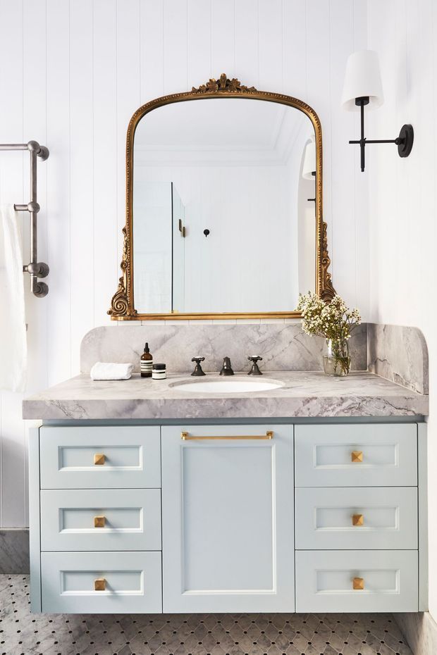 A bright, refined bathroom vanity with a pale blue-grey paneled cabinet, white marble benchtop and integrated backsplash, dark vintage-style tapware, and an ornate antique-gold framed mirror. White vertical wall panelling, black-and-white patterned floor tiles, brass hardware, a wall sconce, and a small floral arrangement complete the elegant traditional fit-out.