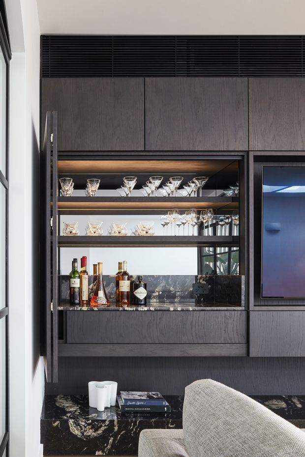 A sophisticated dark-toned built-in bar and entertainment joinery wall, featuring vertically grained dark timber cabinetry, black marble or stone benchtops, mirrored shelving with integrated warm lighting, and neatly displayed glassware and bottles. A television is integrated to the right, with a low marble-topped console and upholstered seating in the foreground. The space has a refined, contemporary feel with strong contrast and luxurious detailing.