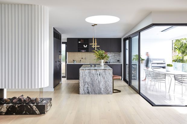Bright contemporary open-plan kitchen with a dark built-in cabinetry wall, textured stone splashback, and a large veined stone island. Pale timber flooring and a sculptural curved white feature wall surround a linear fireplace. Full-height glazed sliding doors open to a furnished outdoor dining terrace, creating a strong indoor-outdoor connection.