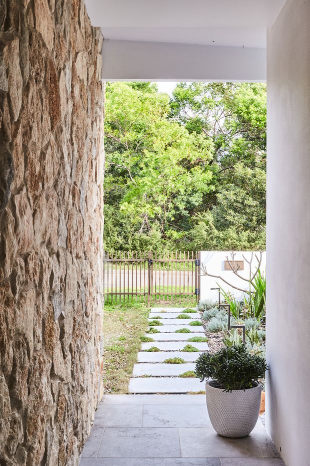A covered entrance passage opens onto a landscaped front garden. The passage has a rugged natural stone feature wall on the left, smooth white rendered walls and ceiling, and large rectangular pale stone or concrete floor tiles. A stepping-stone path leads through low planting toward a timber slat fence and mature trees. A potted succulent sits beside the entry.