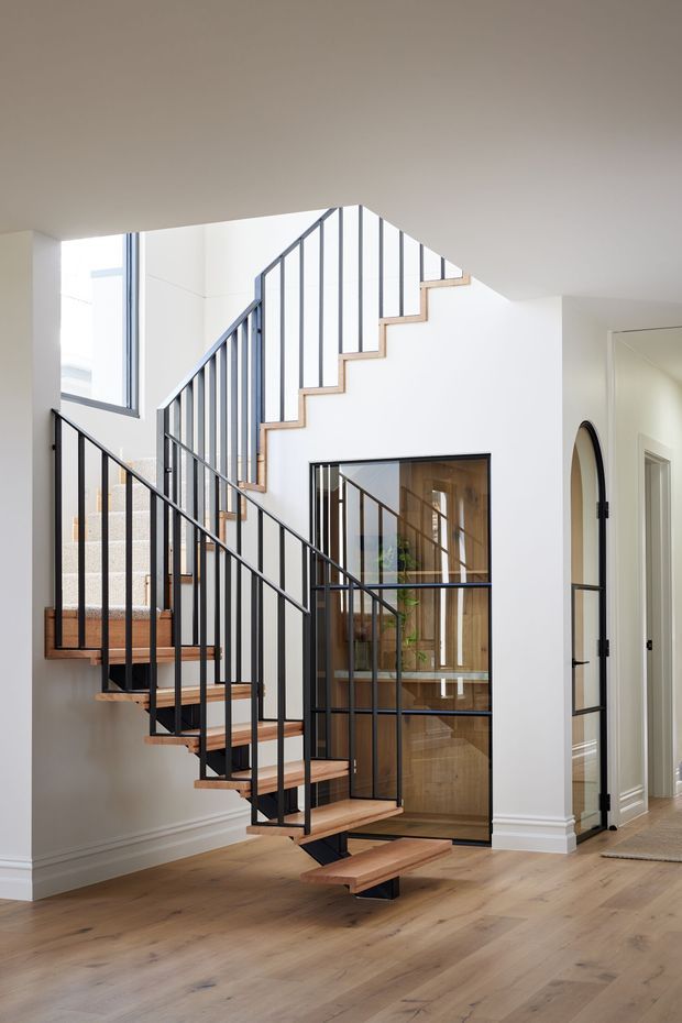 Bright contemporary stairway with floating timber stair treads, slim black metal balustrades, white walls, and warm timber flooring. The stair rises through an open void beside a glazed black-framed partition and doorway, with a glimpse of a timber-lined room beyond.
