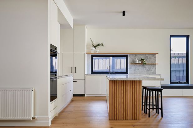 Bright contemporary kitchen with warm timber flooring, white cabinetry, a marble-look backsplash and benchtop, and a compact island clad in vertical timber battens with black bar stools. Black-framed windows, open shelving, integrated appliances, and a white radiator complete the clean, modern interior.