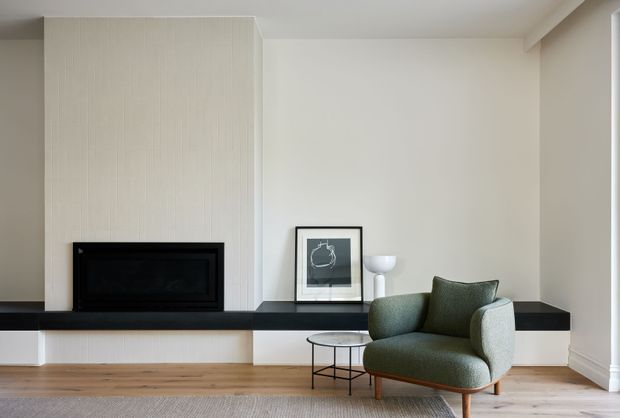 A refined contemporary living room featuring a minimalist fireplace set into a tall vertically textured light wall panel, with a long dark stone hearth or ledge extending across the room. A sculptural green upholstered armchair, small round glass-and-metal side table, framed artwork, and floor lamp add restrained furnishings. Pale timber flooring, neutral walls, and simple architectural detailing create a calm, understated interior.