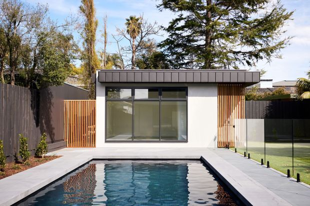 A modern single-storey pool pavilion or backyard extension with a flat roof, white rendered walls, extensive dark-framed glazing, and warm vertical timber screening. It opens onto a rectangular swimming pool surrounded by pale paving, with timber and dark privacy fencing and landscaped planting around the perimeter.