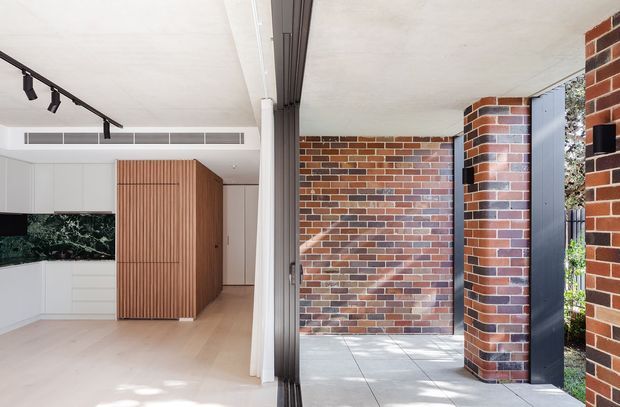 A contemporary residential entry and kitchen viewed through a wide glazed opening. The interior features white flat-panel cabinetry, pale timber flooring, a fluted timber-clad volume, recessed air-conditioning, and black track lighting. Outside, a covered tiled entry court is defined by exposed red and multicoloured brick walls, dark vertical metal or timber screening, and a slender brick column. The composition highlights a strong indoor-outdoor connection and clean, rectilinear detailing.
