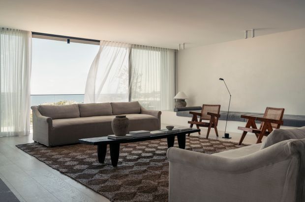 A bright, spacious living room with floor-to-ceiling glazing and sheer curtains overlooking a hazy coastal horizon. The room features a large neutral sectional sofa, sculptural armchair, low black coffee table, patterned area rug, two wooden lounge chairs, a slim floor lamp, and minimal contemporary furnishings.