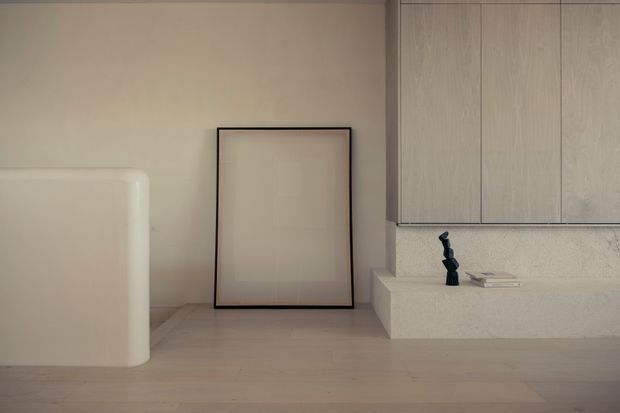 Minimalist interior corner with pale timber flooring, warm off-white plaster walls, a freestanding rounded-edge partition or built-in form, a simple framed artwork leaning against the wall, and light wood-grain cabinetry above a textured stone or concrete ledge. The space is sparsely furnished with a muted, calm palette and soft natural light.