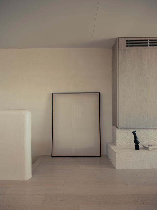 Minimalist interior corner with warm beige plaster walls, pale timber flooring, a softly rounded built-in or freestanding white element, framed neutral artwork leaning against the wall, and light timber cabinetry with integrated ventilation above a raised ledge. A small dark sculptural object and books provide restrained accents.