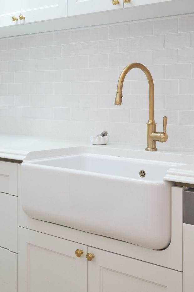 Close-up of a bright, traditional-style kitchen sink area with a large white farmhouse apron-front sink, brushed brass gooseneck tapware, white shaker cabinetry with brass knobs, and pale rectangular tiled splashback.