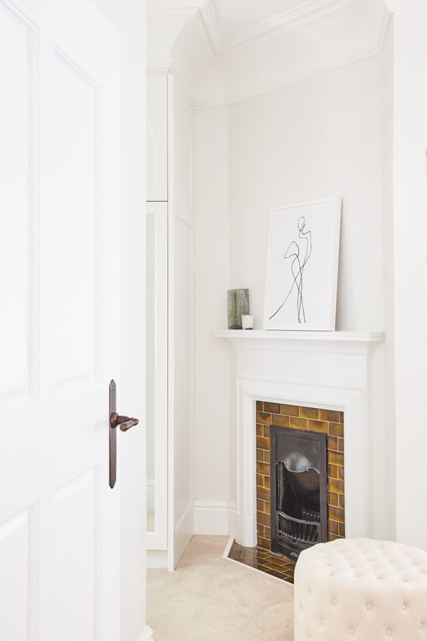 Bright, refined interior corner featuring a traditional white mantel fireplace with a dark metal fire insert and glossy brown-gold rectangular tile surround. White paneled doors, detailed cornices and wall mouldings create a classic architectural character. A slim white built-in cabinet, minimalist line artwork and a cream tufted ottoman complete the space, with pale carpet flooring.