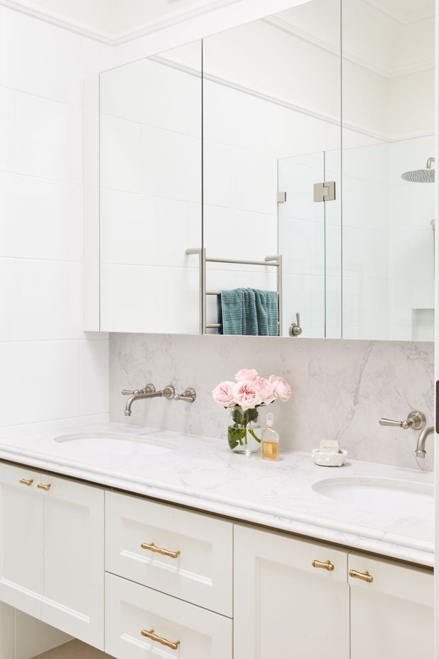 Bright, refined bathroom with a long double vanity, white shaker-style cabinetry, pale marble-look countertop and backsplash, brushed metal wall-mounted taps, and large mirrored cabinetry above. A glass shower enclosure is visible at the right, with folded blue towels and understated floral styling adding soft colour.