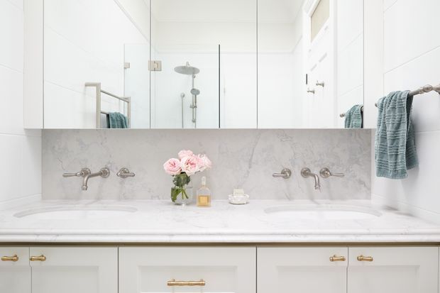 Bright, symmetrical bathroom vanity with a large double basin, white shaker-style cabinetry, pale marble-look benchtop and splashback, wall-mounted brushed metal tapware, and a full-width mirrored cabinet above. A glass shower enclosure and folded blue towels are visible in reflection and at the sides. The space has a clean, classic and refined appearance.