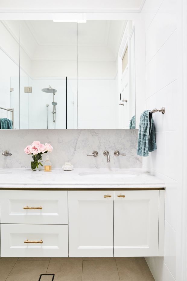 Bright, refined bathroom vanity with a double-basin marble-look benchtop, white shaker-style cabinetry, brushed brass hardware and tapware, large mirrored wall, glass shower enclosure, and white tiled walls. Soft blue towels and pale pink flowers add subtle colour.