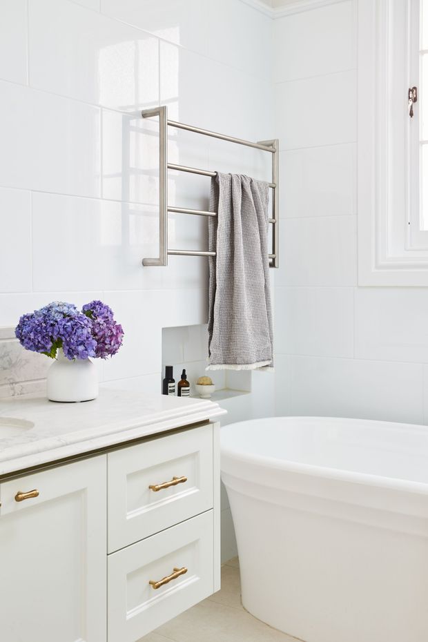 Bright, refined bathroom featuring white tiled walls, a freestanding white bathtub, painted white vanity cabinetry with brushed brass hardware, a pale stone benchtop, and a wall-mounted brushed metal towel rail. A small window provides natural light, while soft grey textiles and purple flowers add subtle colour.