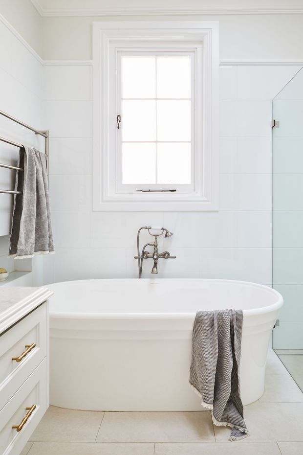 Bright, white bathroom featuring a freestanding bathtub beneath a multi-pane window, a traditional chrome wall-mounted faucet, white tiled walls, pale tiled flooring, a glass shower enclosure, heated towel rail, and white built-in vanity cabinetry with brass handles. The space has a clean, classic and understated appearance.