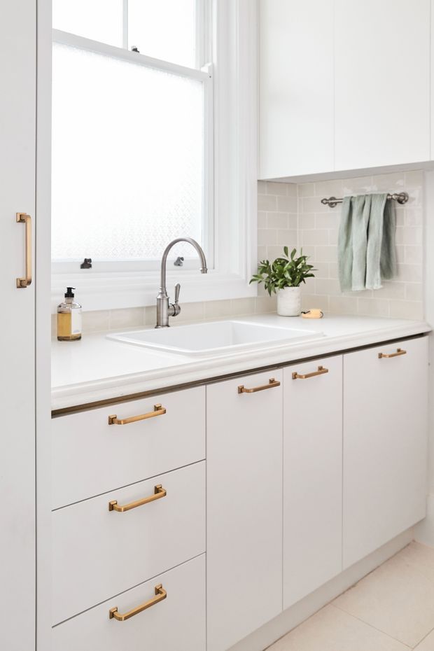Bright, compact bathroom vanity with white cabinetry and a broad white countertop incorporating a rectangular undermount sink and brushed metal gooseneck tap. Pale subway-tile splashback, frosted window, white upper cabinetry, and warm brass cabinet pulls create a clean, fresh fit-out. A small potted plant and folded towels add restrained styling.