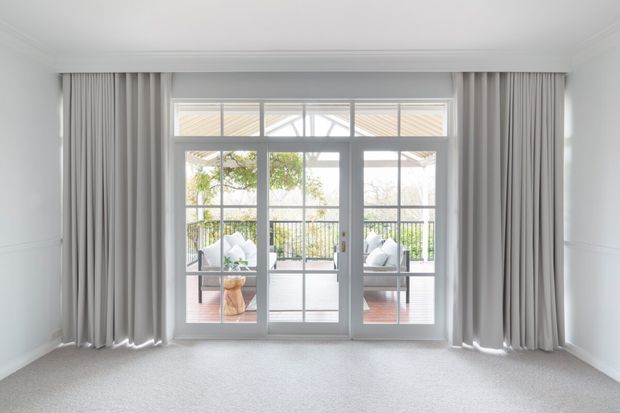 Bright, empty interior room with pale carpet, white walls, restrained cornice and wall panelling, and full-height grey curtains framing large white-framed glazed French doors. The doors open to a furnished terrace or balcony with outdoor seating and greenery beyond.