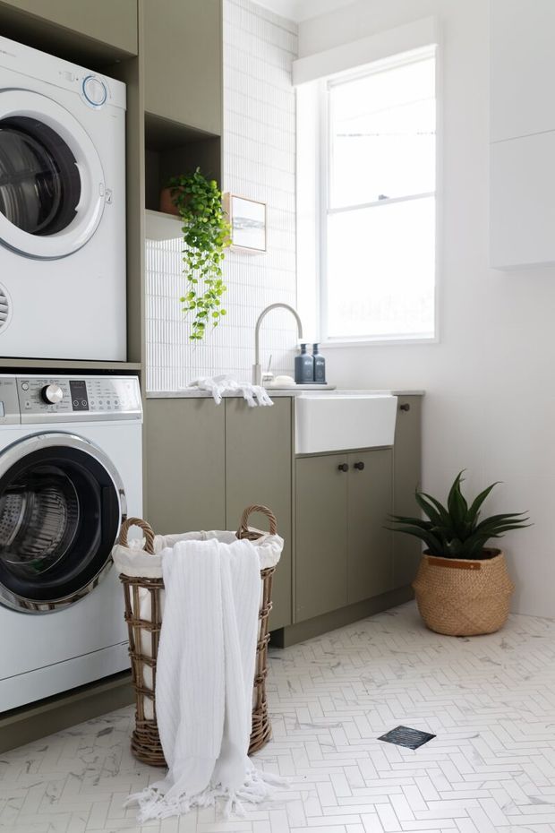 Bright, compact laundry room with stacked front-loading washer and dryer integrated into muted green-grey cabinetry. A white utility sink with brushed metal gooseneck tap sits beneath a large window, with white vertically textured wall tiles, white herringbone floor tiles, open shelving, woven laundry baskets, and a potted plant. The overall look is clean, practical, and softly contemporary.