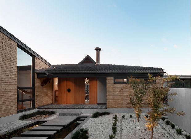 Exterior view of a low, single-storey brick residence with a dark tiled pitched roof, prominent timber entrance door, tall glazing, and a landscaped gravel courtyard. A chimney rises from the roof, while stepping stones cross a shallow linear water feature or dark drainage channel toward the recessed entry.