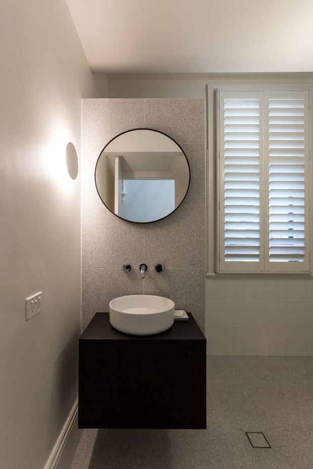 A compact, refined bathroom featuring a floating dark timber vanity with a white vessel basin, wall-mounted taps, a round black-framed mirror, pale stone-look wall tiles, and a grey tiled floor. A shuttered window provides natural light, while soft warm lighting creates a calm, understated atmosphere.
