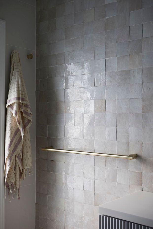 A close-up view of a refined bathroom shower or wet-area wall, finished in irregular handmade-looking pale grey and off-white square tiles with varied texture and sheen. A slim brass-gold grab rail runs horizontally across the tiled wall. At left, a striped neutral textile hangs from a wall hook, while the lower right shows the edge of a white vanity with dark vertical slatted detailing. The mood is calm, tactile, and understated.