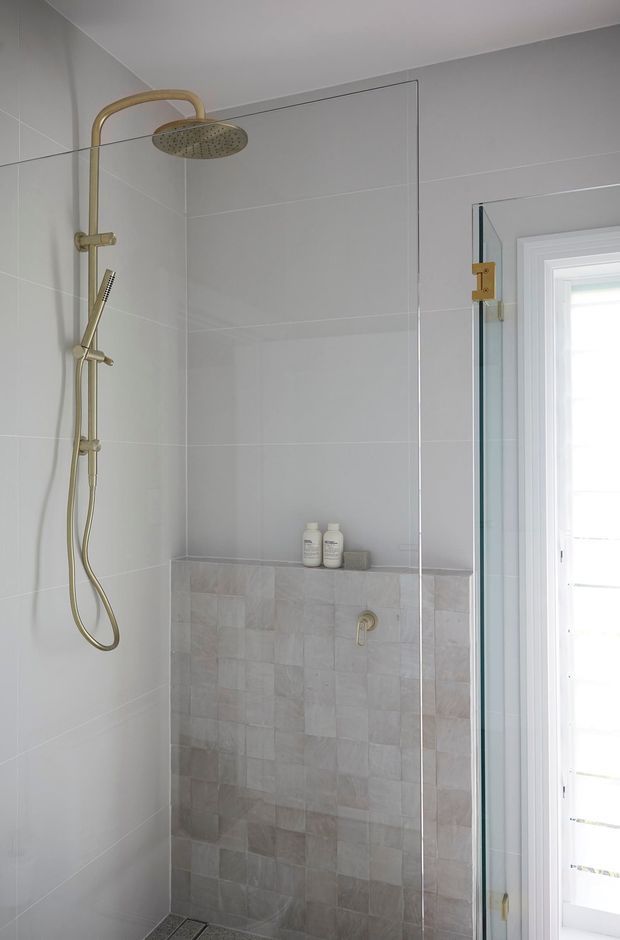 A contemporary walk-in shower enclosed by clear glass, with large pale grey wall tiles, a contrasting lower wall section of textured square tiles, and brushed brass shower fittings. A white-framed window provides natural light.