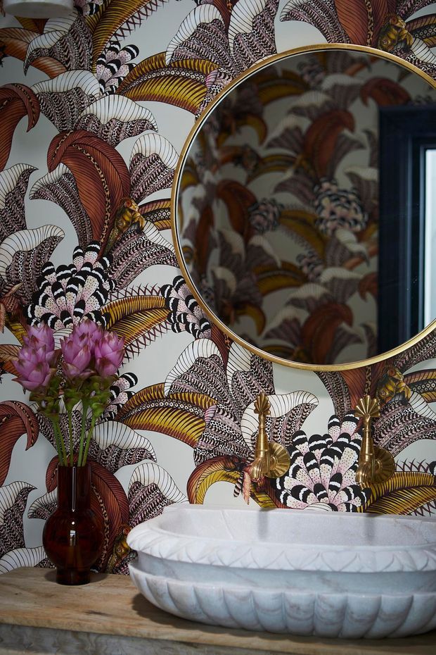 A richly styled bathroom vignette featuring a white scalloped vessel basin on a pale stone or timber-toned vanity, paired with ornate brass wall-mounted taps. The wall is covered in dramatic botanical wallpaper with fan-shaped leaves in burgundy, mustard, black, cream and blush tones. A round brass-framed mirror reflects the patterned wall, while a dark glass vase of pink flowers adds a decorative accent.