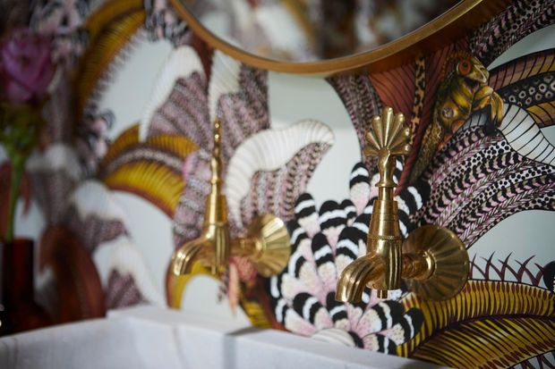 Close-up of an ornate bathroom vanity or wash area featuring a highly decorative patterned wall surface with stylised birds, feathers and botanical motifs. Two polished brass/gold wall-mounted tap fixtures are visible above a white basin or countertop, with a curved framed mirror or reflective surface above.