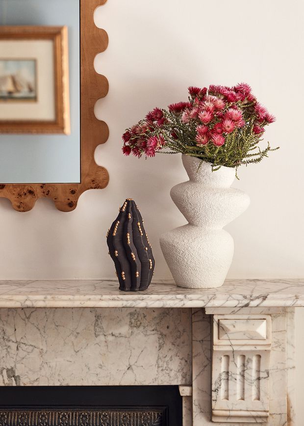 Close-up interior detail of a marble fireplace mantel with veined stone, ornate carved detailing, and a dark decorative firebox surround. The mantel is styled with a sculptural white textured vase holding pink flowers, a dark ribbed decorative vessel, and a large organic-framed mirror with a timber frame. The composition has a refined, vintage-inspired decorative character.