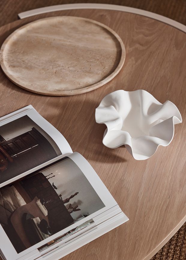 Styled tabletop vignette featuring a warm timber surface, a shallow round stone or travertine tray, a sculptural white organically folded bowl, and an open design magazine. The composition is softly lit and presented in a refined, warm neutral palette.