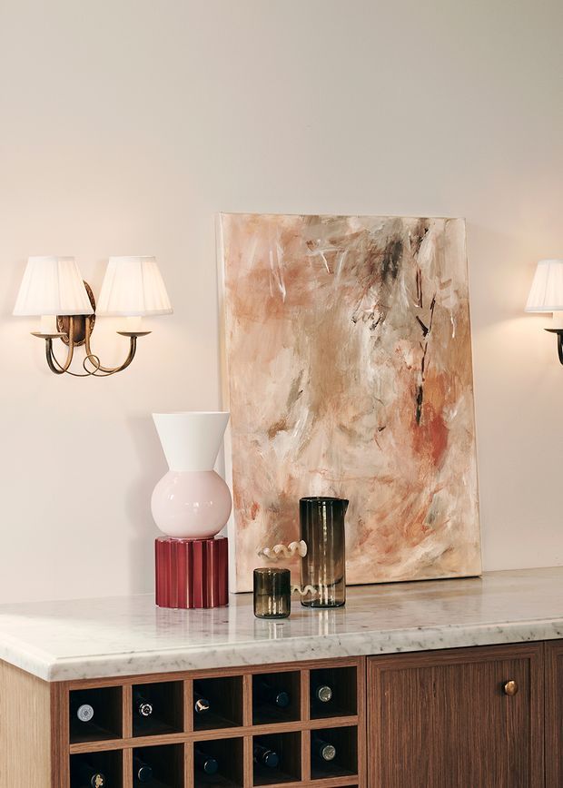 A refined interior vignette featuring a marble-topped timber sideboard with open wine storage and paneled cabinetry. A large abstract painting leans against a warm neutral wall, complemented by sculptural vases, glass vessels, and traditional wall sconces with fabric shades.