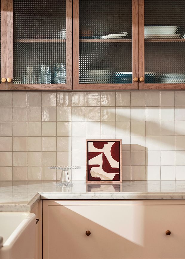 Warm, softly lit kitchen detail featuring natural timber upper cabinetry with textured glass doors, glossy cream square-tile splashback, pale stone benchtop, and blush-beige lower cabinetry. A small framed abstract artwork and glassware add decorative accents, with strong sunlight casting geometric shadows across the joinery.