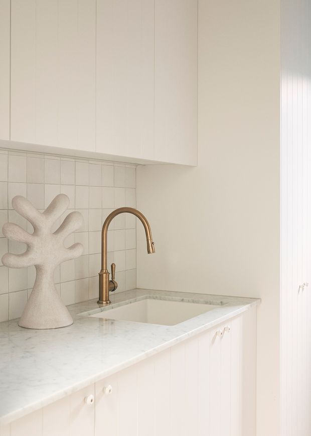 A close-up of a light, minimalist kitchen work area featuring white vertical-panel cabinetry, a pale marble countertop, a white tiled backsplash, an undermount sink, and a curved brushed brass tap. A sculptural ceramic object sits on the counter, while vertical blinds are visible at the right edge.