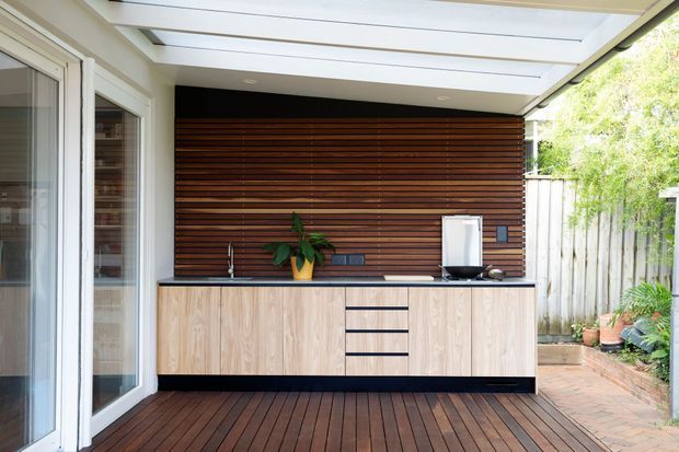 A compact covered outdoor kitchen or entertaining area with light timber cabinetry, a dark benchtop, horizontal dark timber wall slats, and a timber deck. A small sink, cooktop or appliance area, and countertop appliances are visible, with a translucent roof providing shelter. Glazed doors open to the left, while a paved garden boundary is visible to the right.