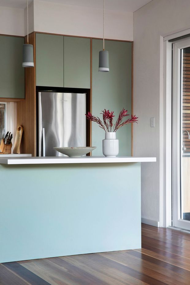 A compact contemporary kitchen viewed from the living area, featuring a pale sage-green island and matching upper cabinetry framed in warm timber. The kitchen includes a stainless-steel refrigerator, white countertop, pendant lights, and dark timber flooring. A glazed door or window provides bright natural light at right, while simple ceramic vessels and understated floral branches add restrained styling.