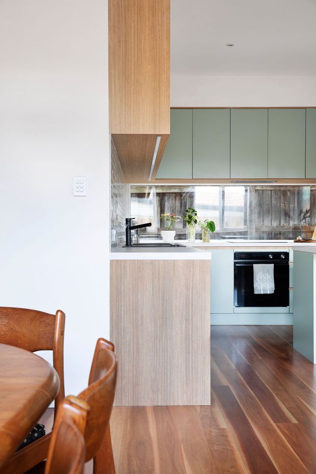 A bright, compact contemporary kitchen viewed from an adjoining dining area. The kitchen features muted sage-green flat-panel cabinetry, light timber cabinetry and island cladding, white benchtops, a dark tap and oven, a tiled backsplash, and warm timber flooring. A timber dining table and chairs are visible in the foreground.